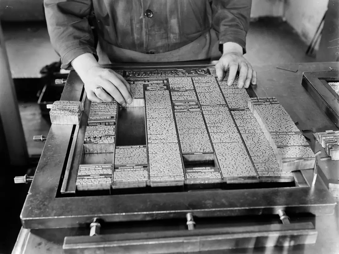 A newspaper make-up man's hands arranging lead type for a front page in a steel frame.